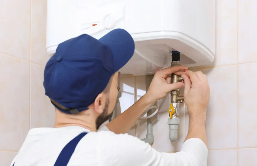 Plumber wearing a blue cap tightening pipes under a wall-mounted water heater.