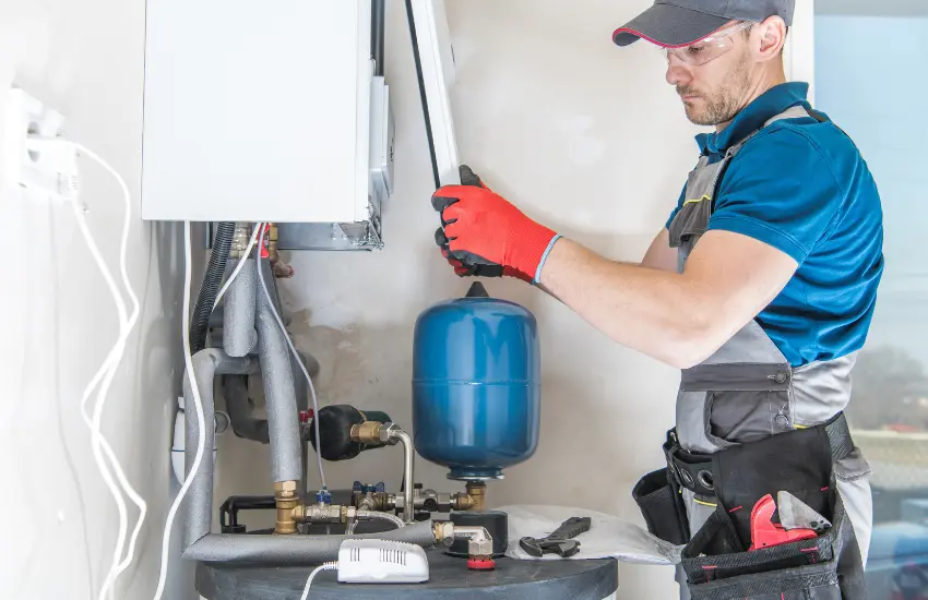 Worker in blue uniform with red gloves servicing a home boiler, adjusting a valve beside a blue expansion tank.